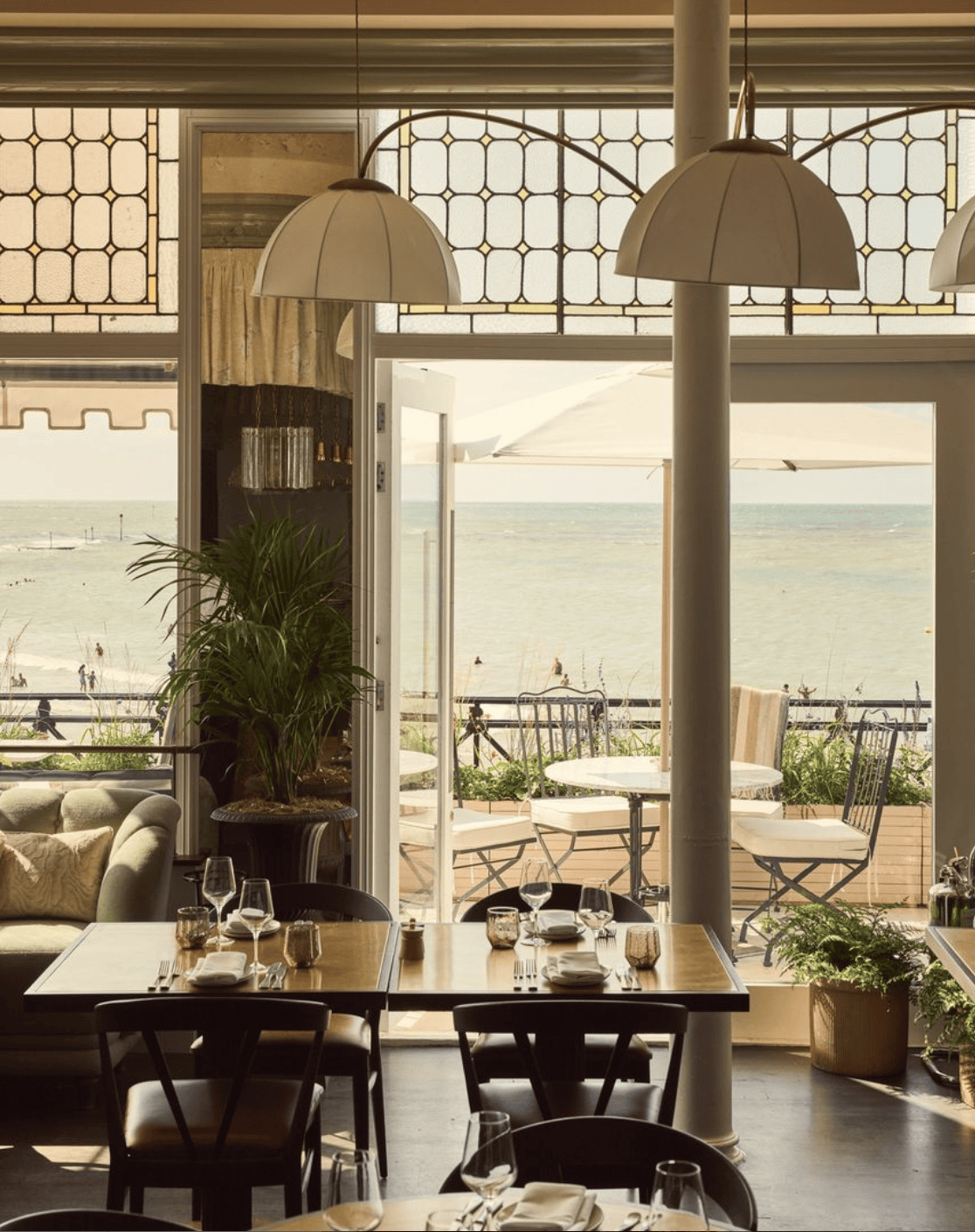 Restaurant interior with ocean view. Tables set with place settings, chairs, and hanging lamps.