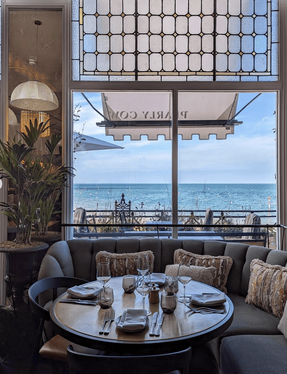 Restaurant seating with ocean view. Round table set, plush seating, large window with stained glass and awning.