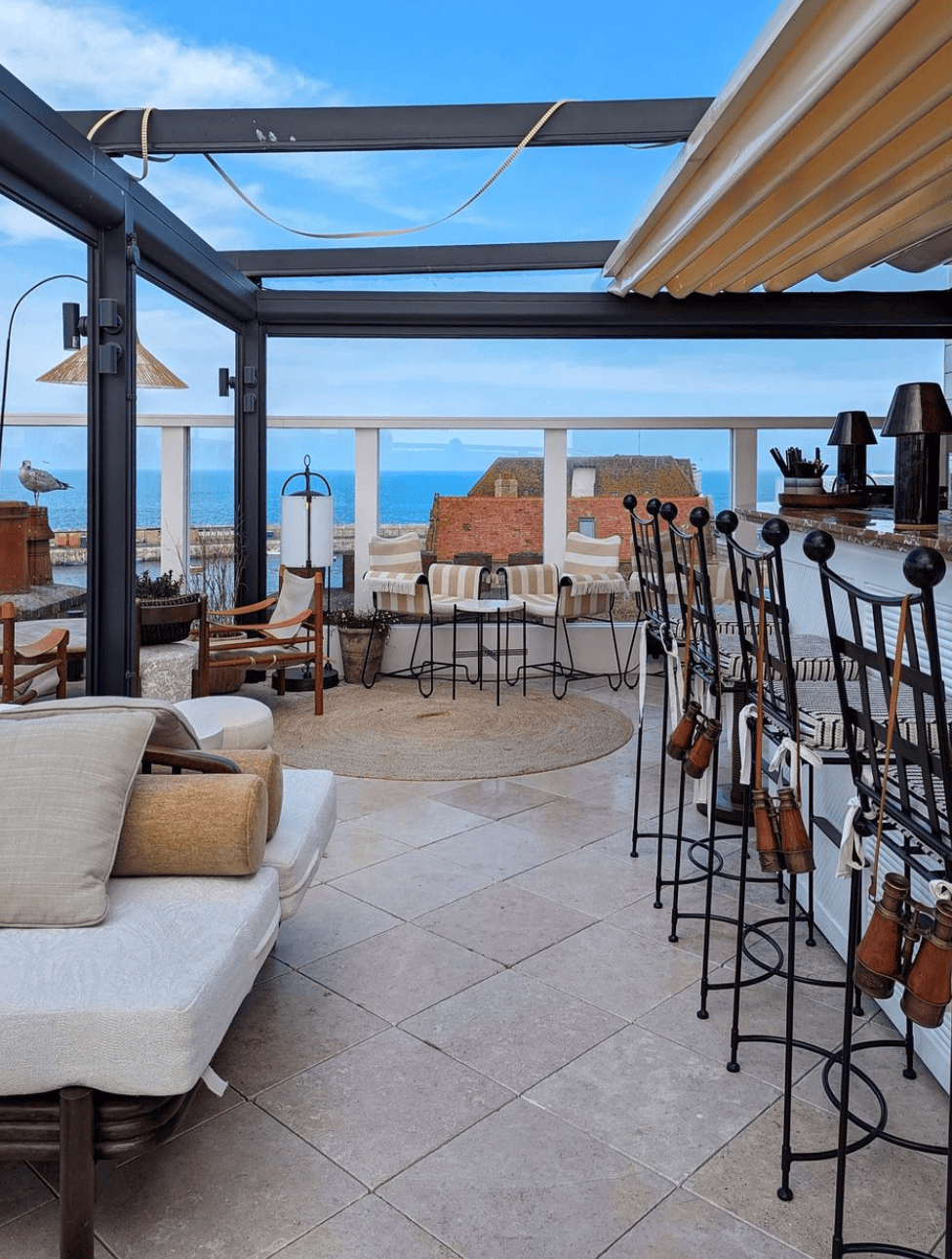 Rooftop bar with cushioned seating, stools, and ocean view under a blue sky.