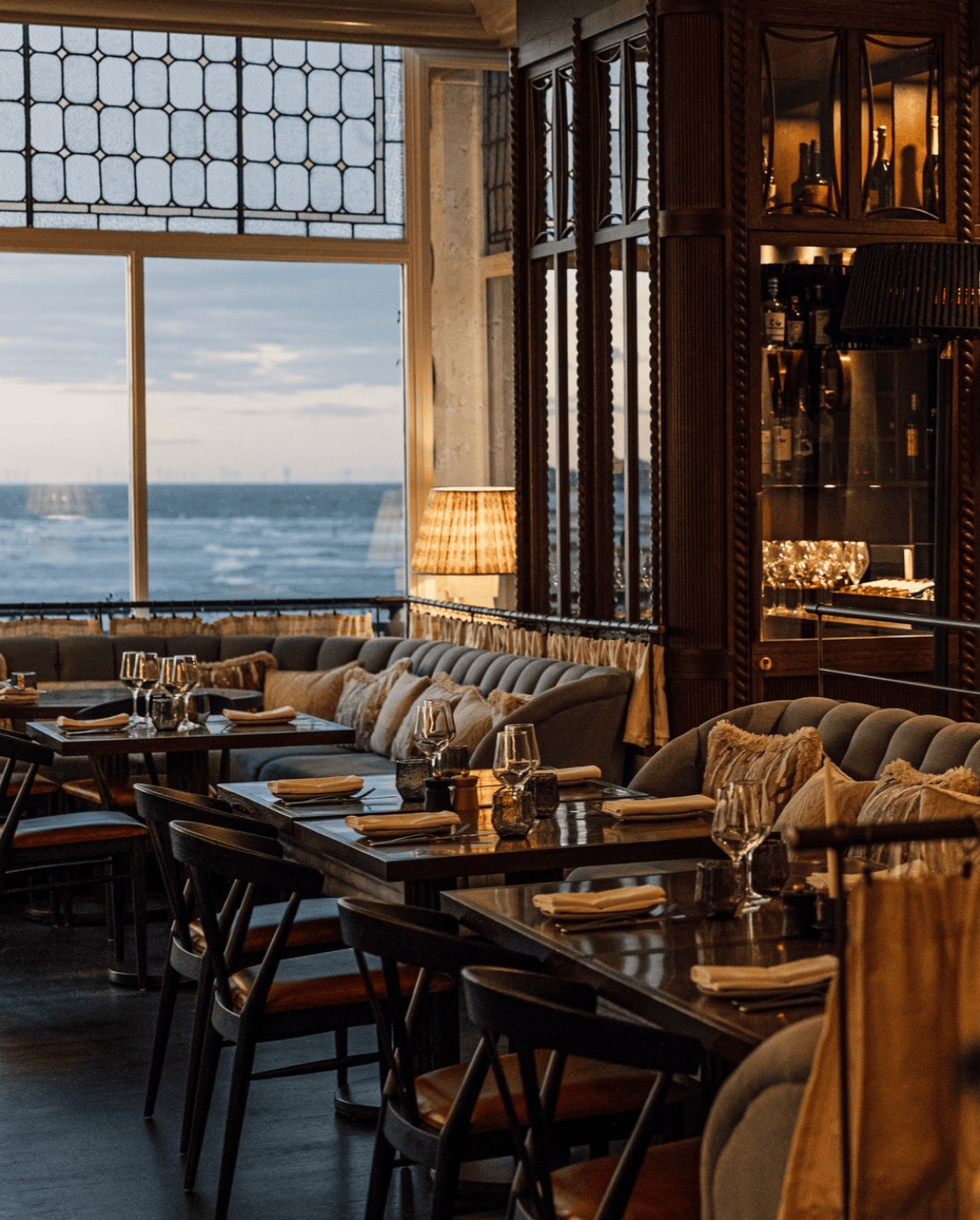 Restaurant interior with tables set for dining and ocean view. Dark wood, plush seating, and large windows.
