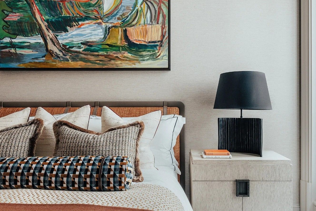 Bedroom with art, lamp, and woven headboard, featuring patterned pillows and a textured nightstand.