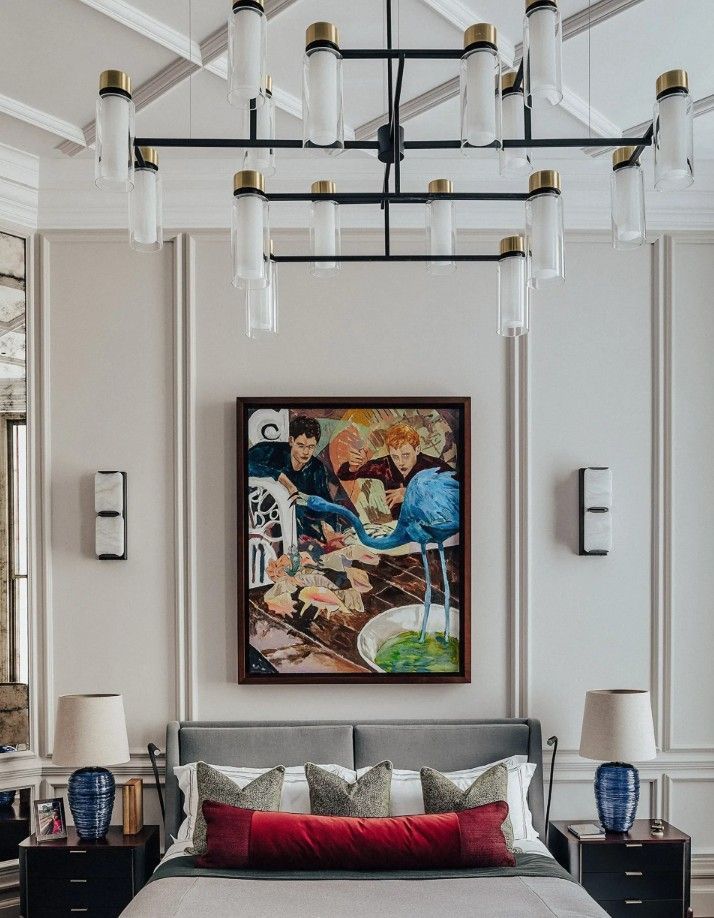 Bedroom with a painting above the bed, a chandelier, two lamps, and light-colored walls with molding.