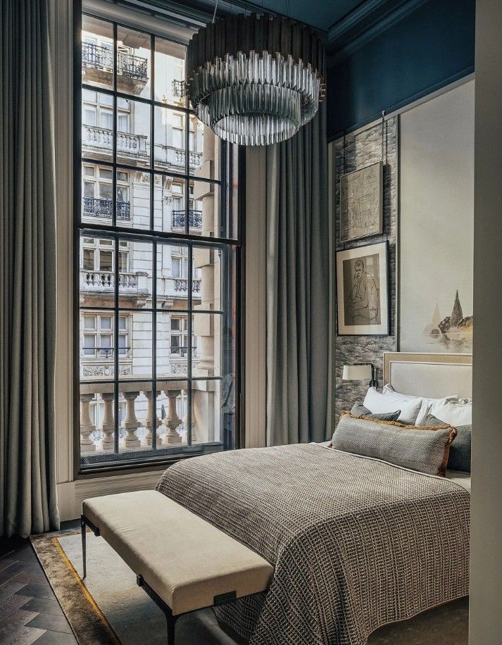 Bedroom with large window overlooking a city street. Gray bed, bench, curtains, and a chandelier.