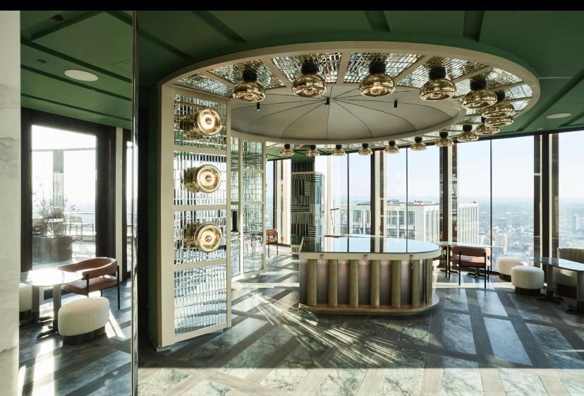 Stylish bar with round counter, surrounded by windows offering city views, in a green and beige interior.