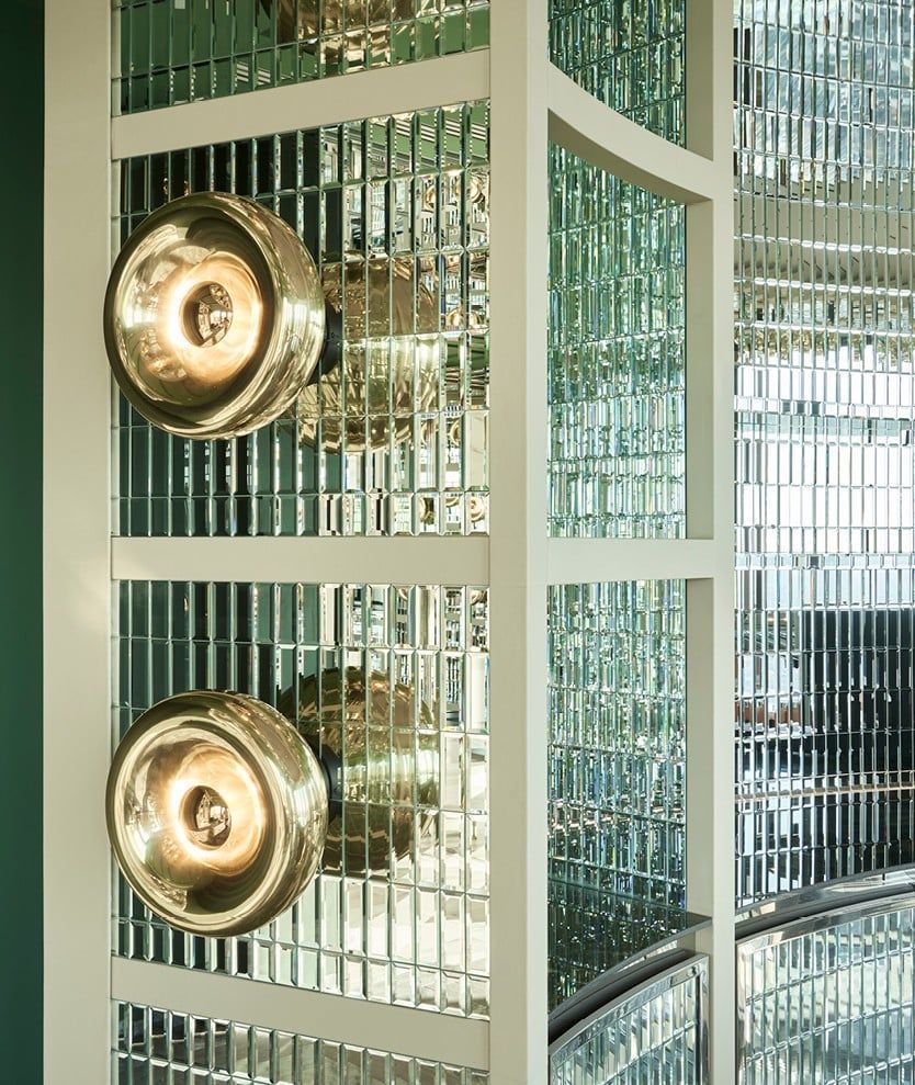 Two gold sconces on a white frame with a mirrored tile wall, reflecting light and details.