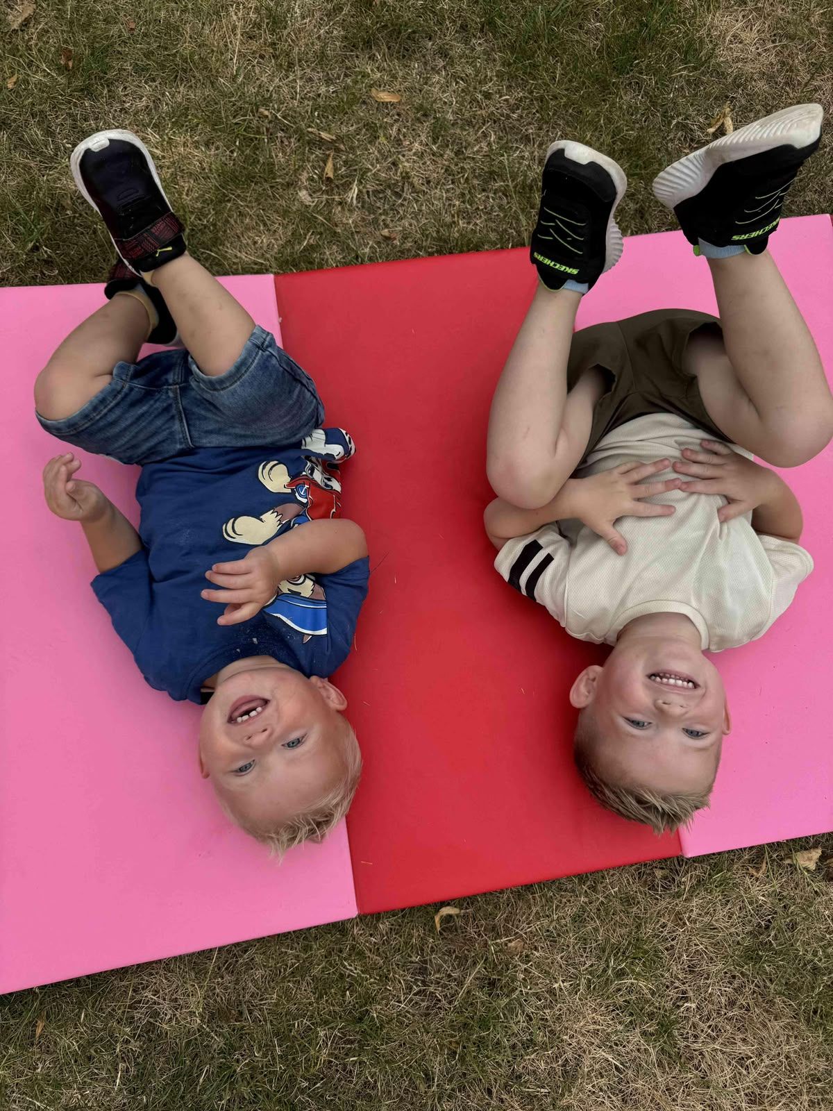 Twee kinderen liggen lachend op de camera op een roze-rood kleed op het gras