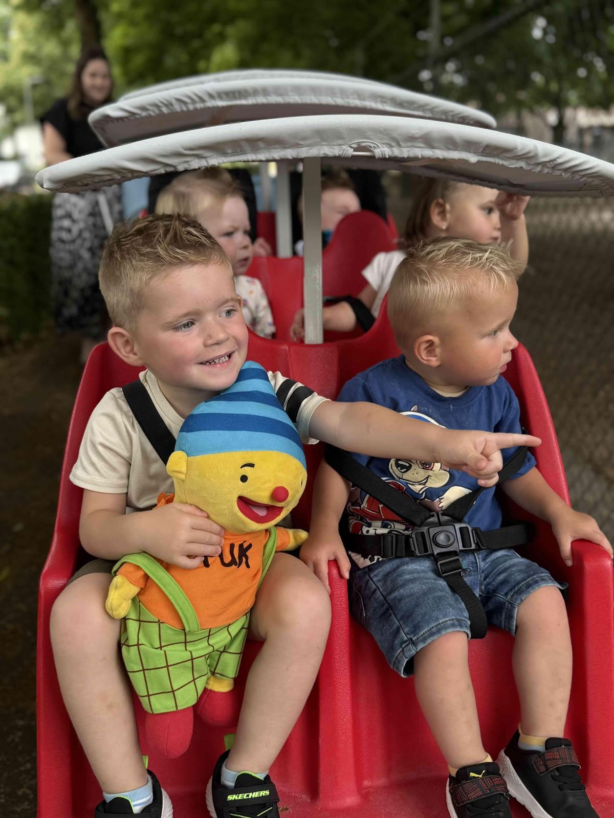 Drie kinderen zitten in een rode kinderwagen. Eén van hen houdt een knuffel vast en glimlacht buiten.