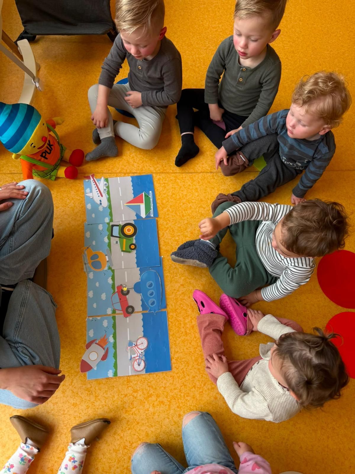 Kinderen zitten op een geel tapijt rond een kleurrijk bordspel en kijken en spelen samen
