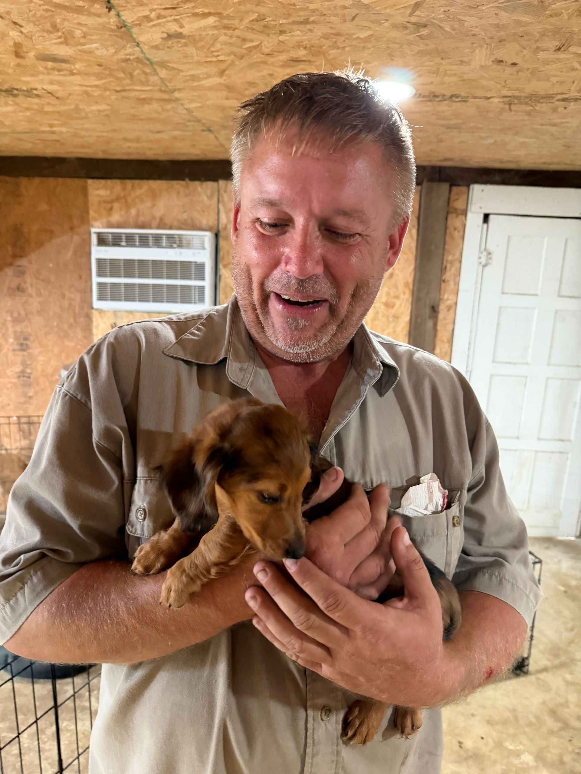 Man holding a small brown puppy indoors, smiling at it.
