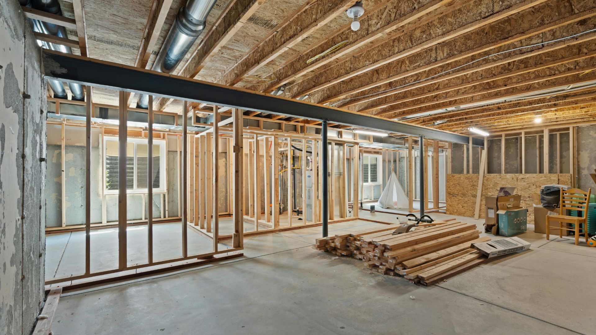 Basement renovation with exposed wooden framing, concrete floor, and overhead ductwork. Construction materials visible.