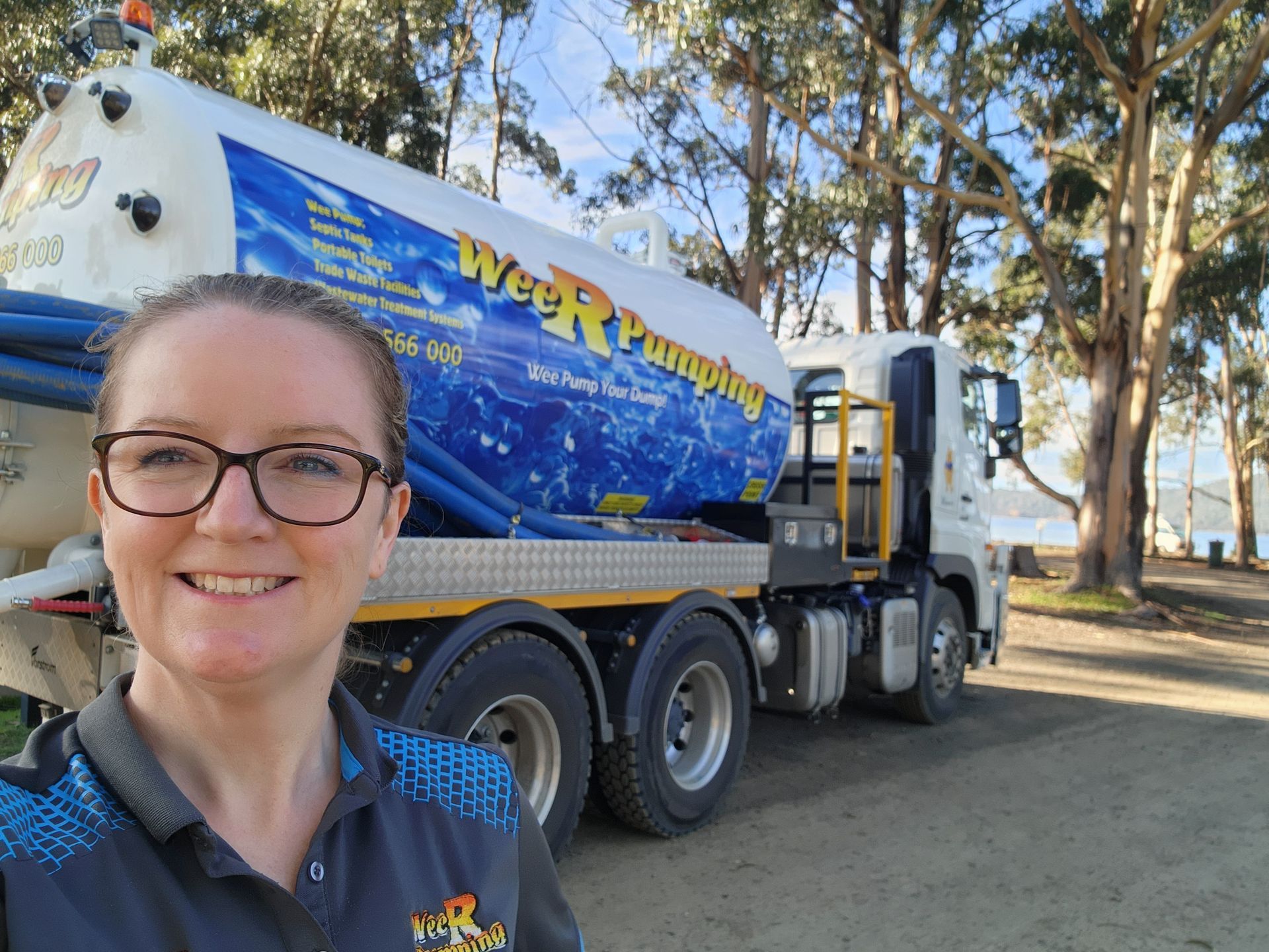 Smiling person in glasses stands beside a blue tanker truck in a wooded outdoor area — Wee R Pumping In Glenorchy, TAS