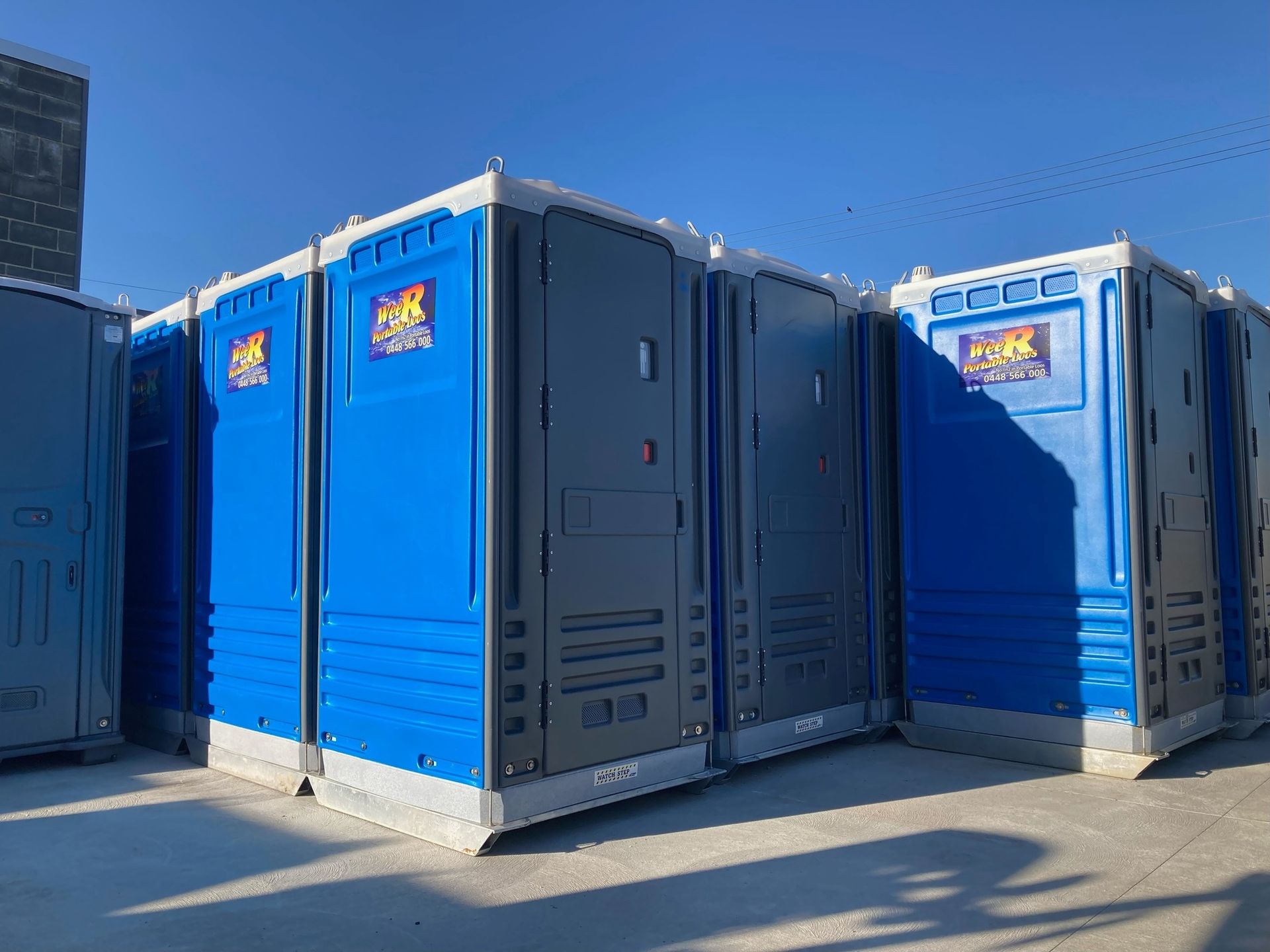 Blue Portable Toilets Lined Up Outdoors in Bright Sunlight Against a Blue Wall — Wee R Pumping In Glenorchy, TAS