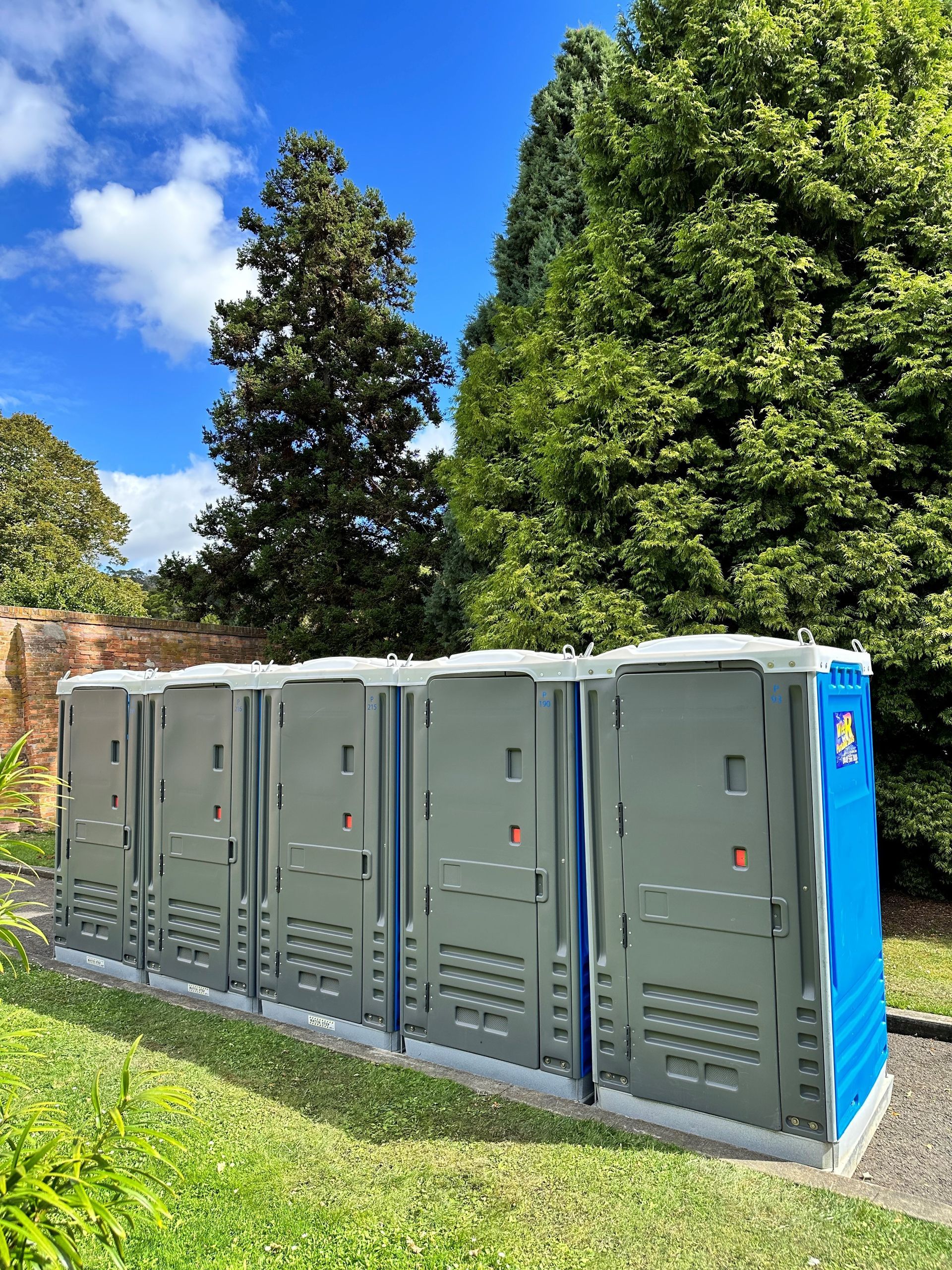 Row of gray portable toilets outdoors beside a brick wall, with trees and blue sky in the background — Wee R Pumping In Glenorchy, TAS