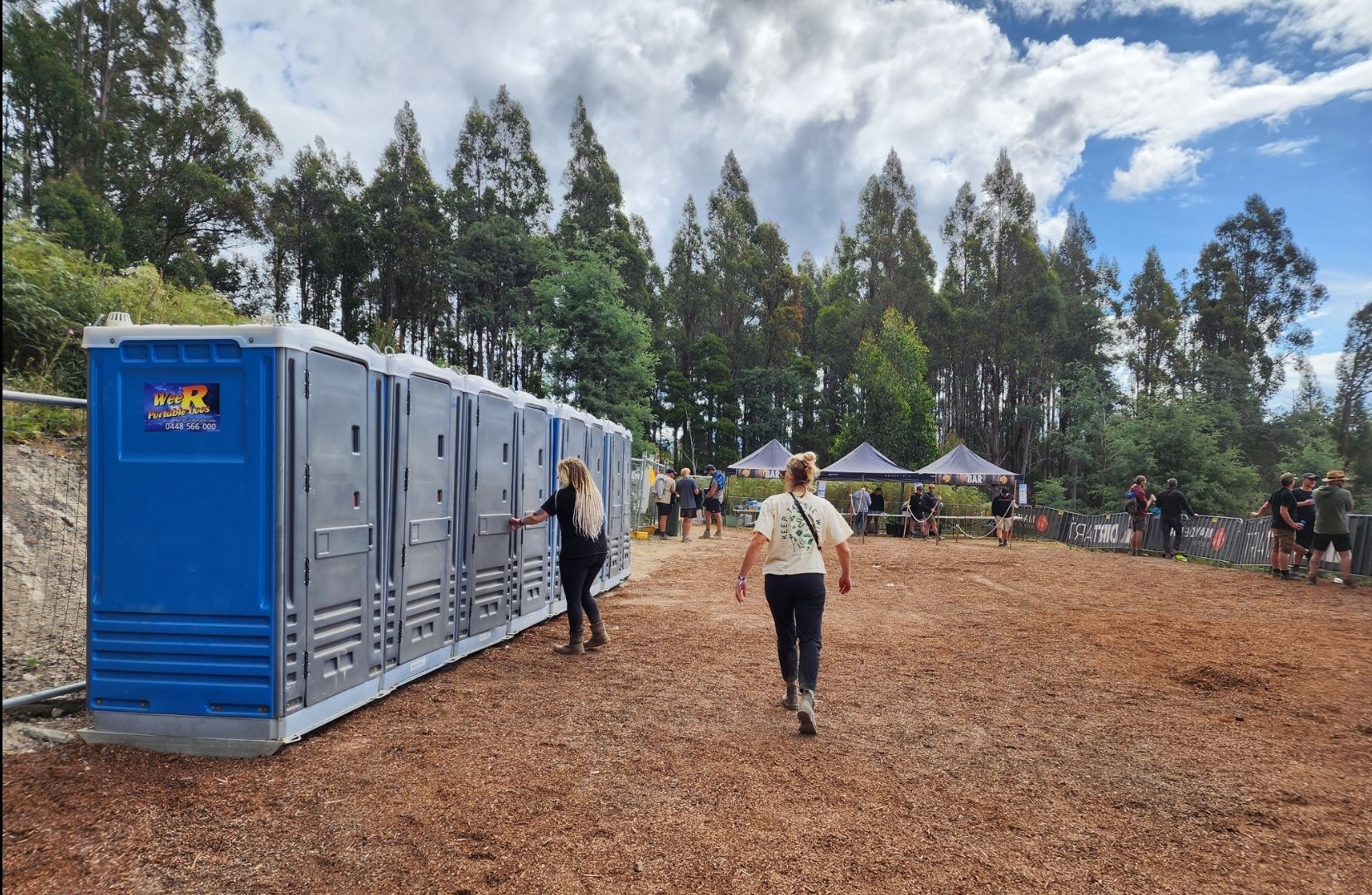 Portable toilets lined up at an outdoor event site with people walking on a dirt path under cloudy skies — Wee R Pumping In Glenorchy, TAS