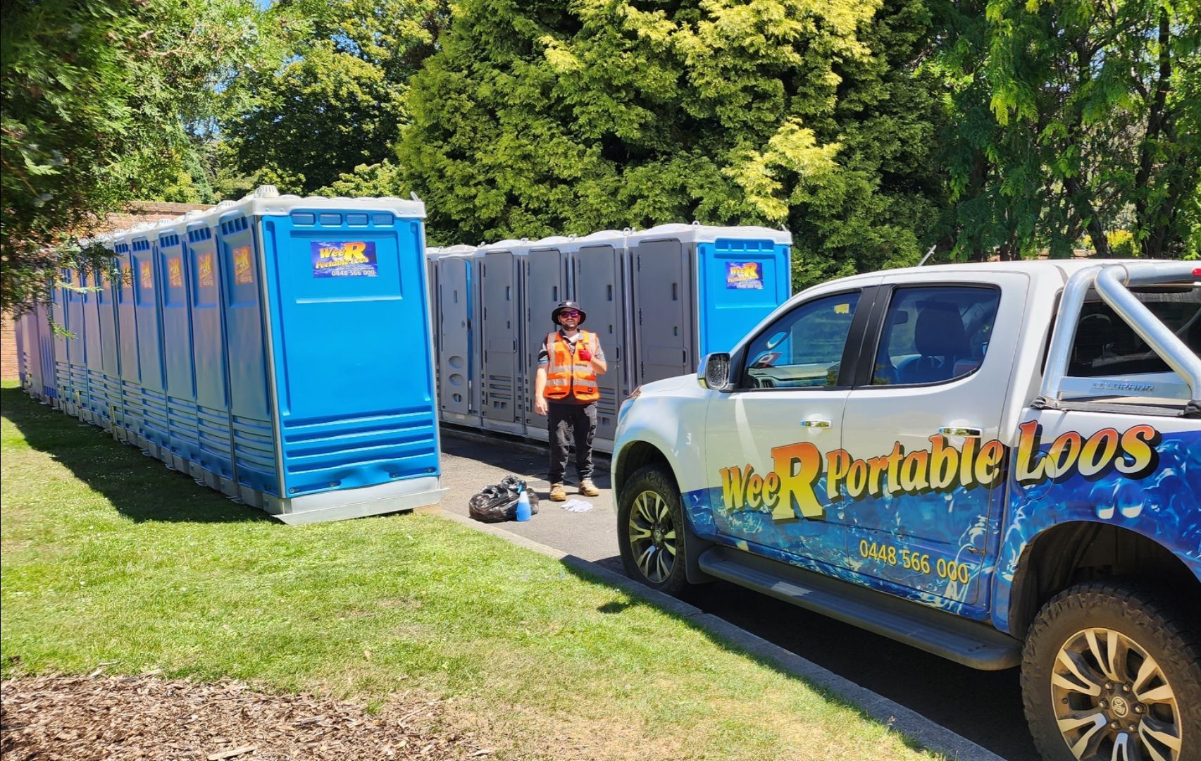 Blue portable toilets lined up on grass beside a work truck and a worker in an orange vest.— Wee R Pumping In Glenorchy, TAS