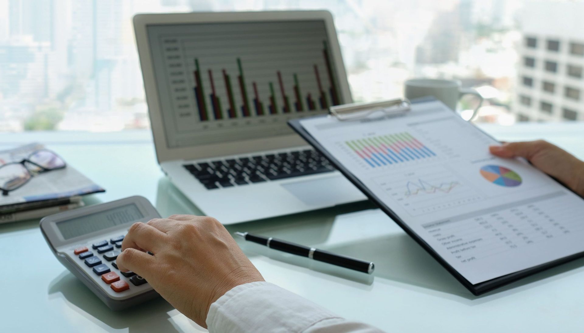 Person calculating numbers with a calculator, reviewing charts on a clipboard and laptop. Office setting with cityscape view. Person calculating numbers with a calculator, reviewing charts on a clipboard and laptop. Office setting with cityscape view.