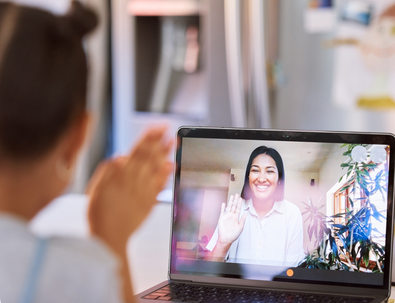 A young girl waves at a smiling woman on a laptop screen, inside a home with a fridge in the background. A young girl waves at a smiling woman on a laptop screen, inside a home with a fridge in the background.