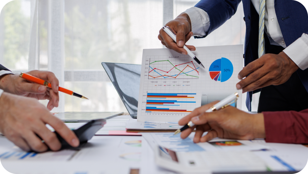 People in business attire reviewing charts and graphs during a meeting at a table, using pens to point at data. People in business attire reviewing charts and graphs during a meeting at a table, using pens to point at data.