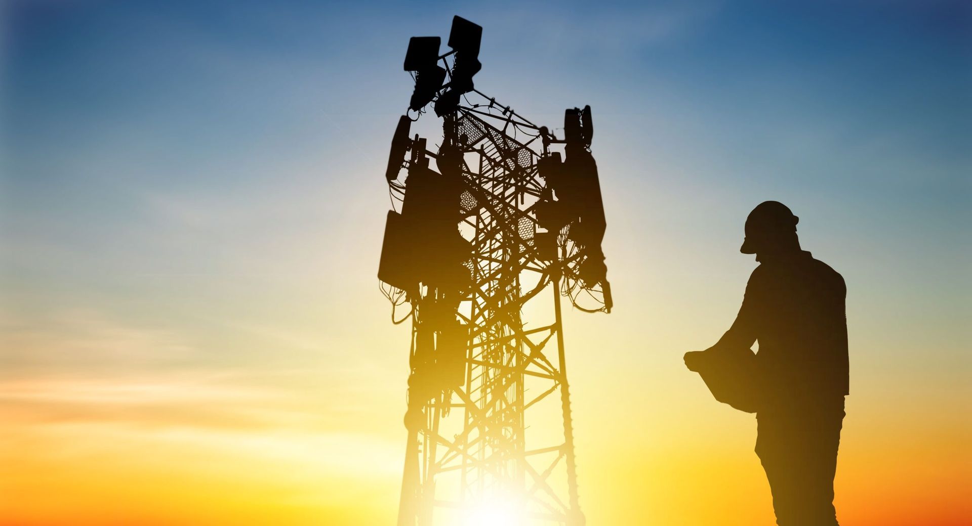 Silhouette of a worker in a hard hat inspecting a communications tower against a sunset. Silhouette of a worker in a hard hat inspecting a communications tower against a sunset.
