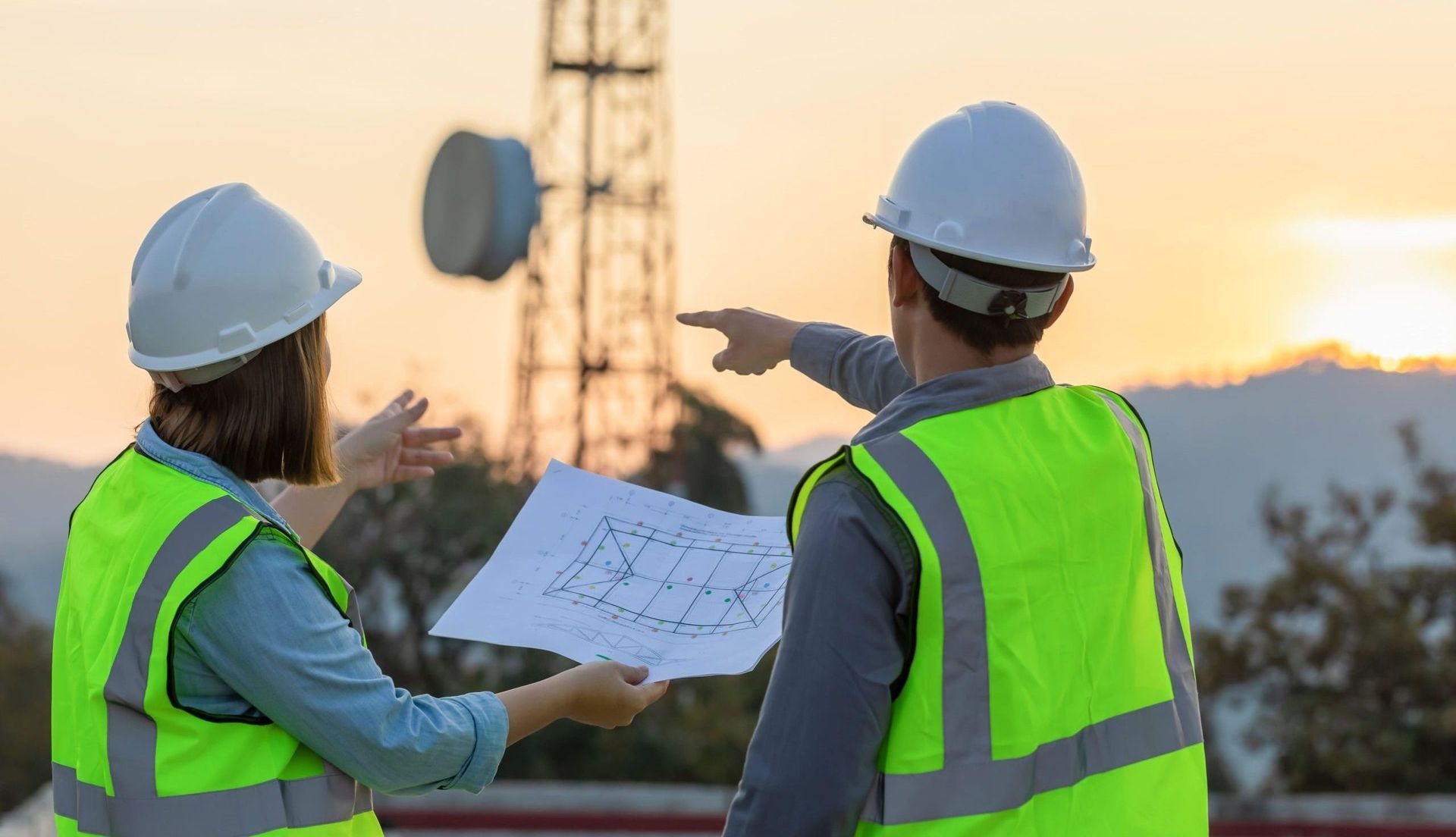 Two engineers in safety vests and hard hats examine a blueprint near a telecommunications tower at sunset. One points. Two engineers in safety vests and hard hats examine a blueprint near a telecommunications tower at sunset. One points.