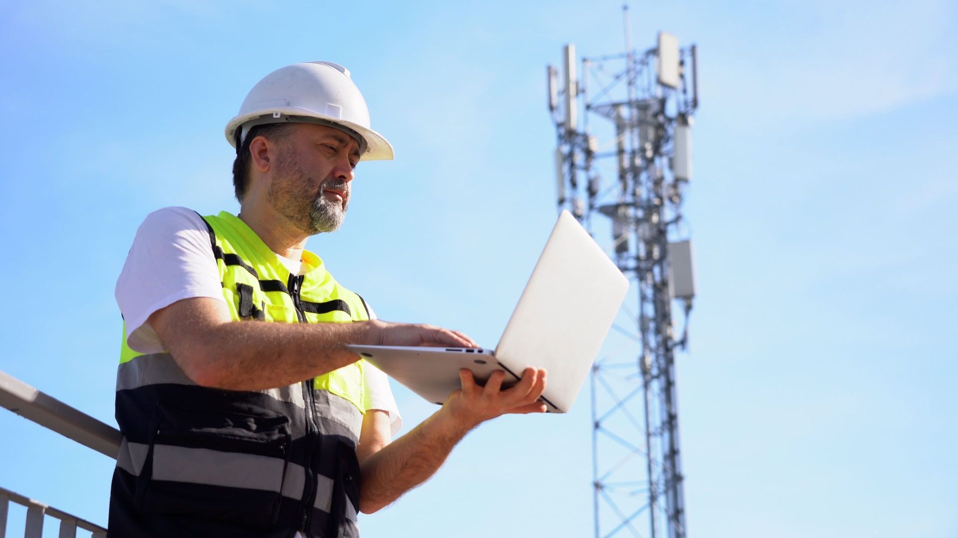 A telecom worker in a hard hat and vest using a laptop, with a cell tower in the background against a blue sky. A telecom worker in a hard hat and vest using a laptop, with a cell tower in the background against a blue sky.