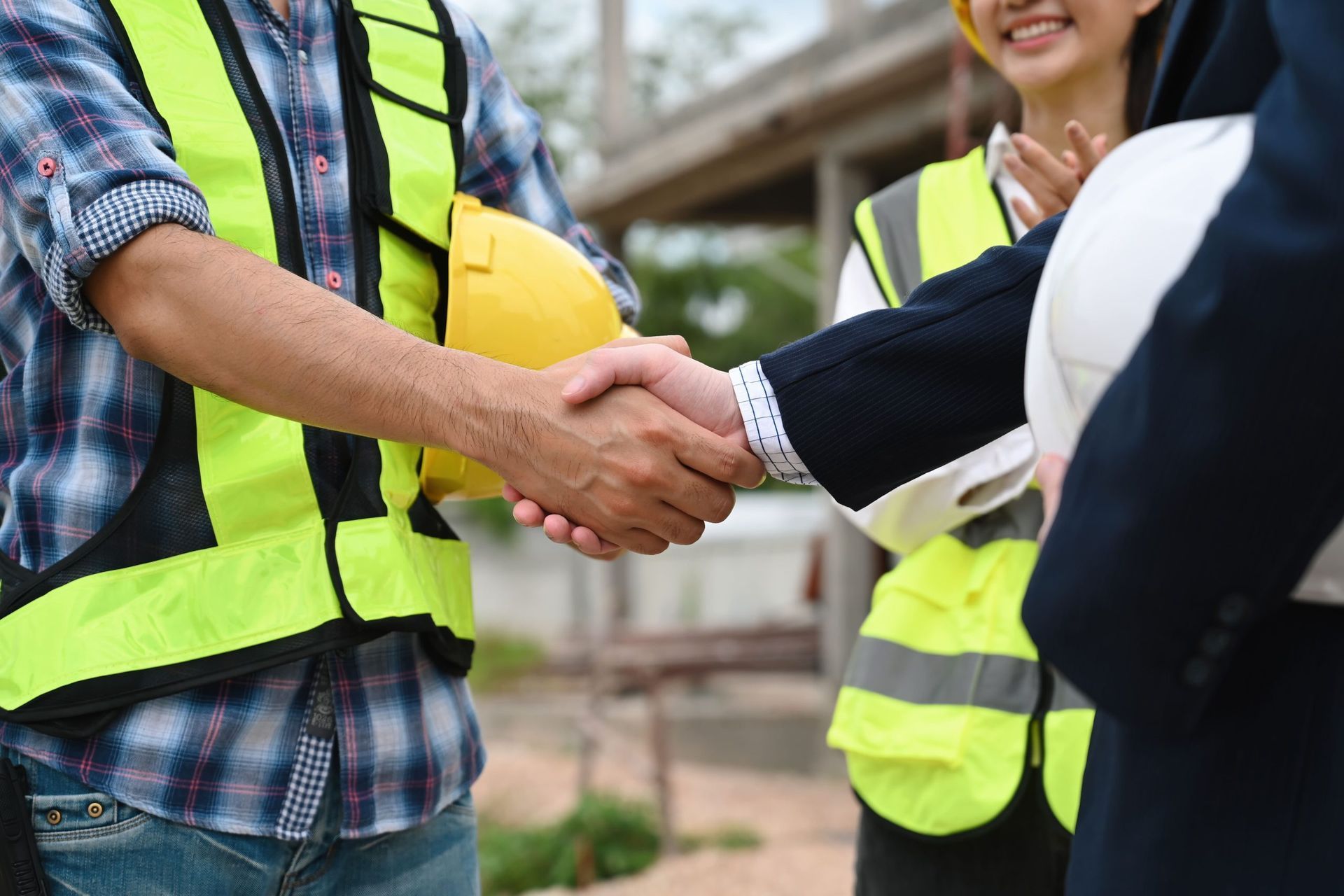 Construction workers shaking hands, signifying agreement. A person wears a safety vest and holds a yellow hard hat. Other workers in the background. Construction workers shaking hands, signifying agreement. A person wears a safety vest and holds a yellow hard hat. Other workers in the background.
