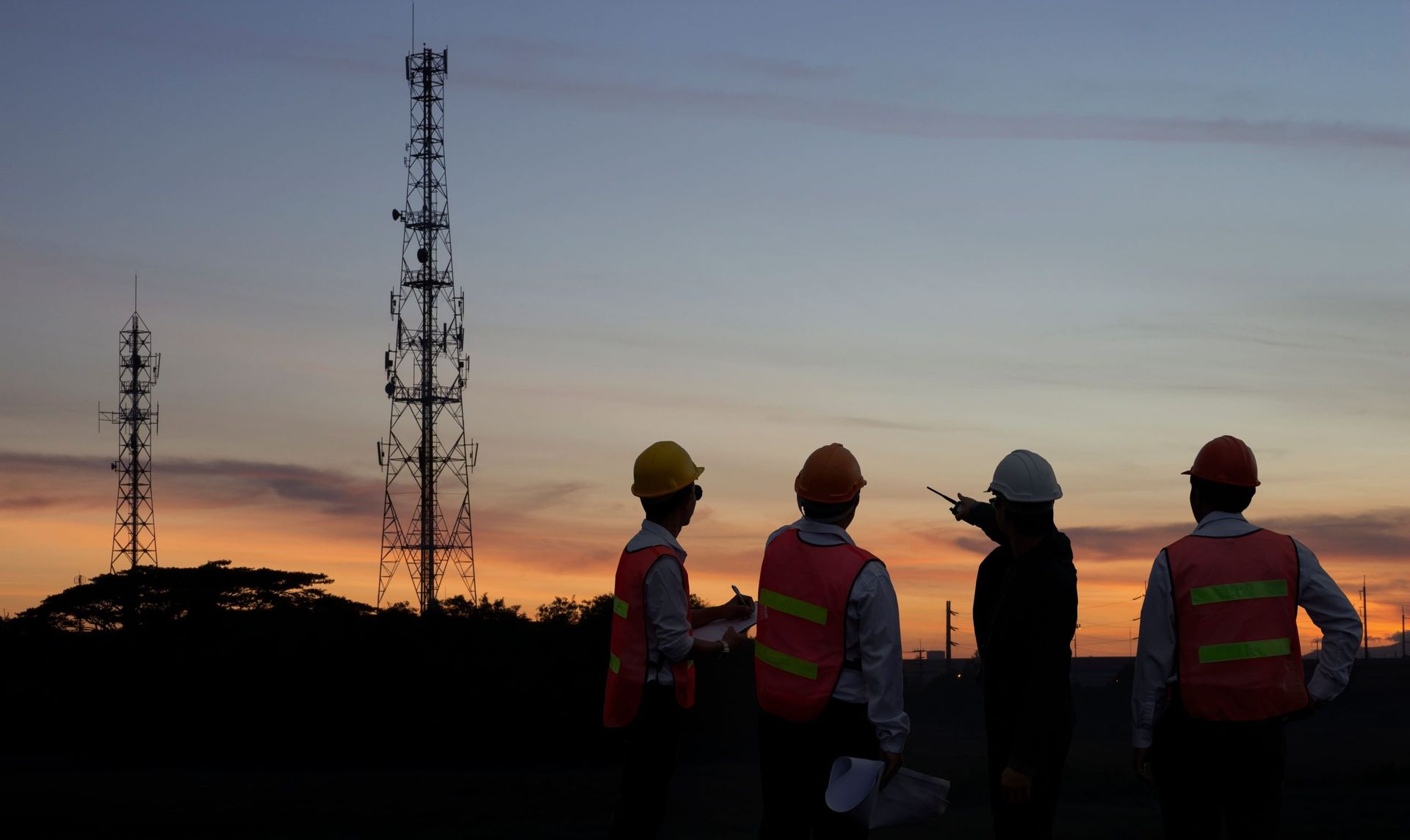 Silhouette of engineers in safety vests and hard hats, inspecting cell towers at sunset. Silhouette of engineers in safety vests and hard hats, inspecting cell towers at sunset.