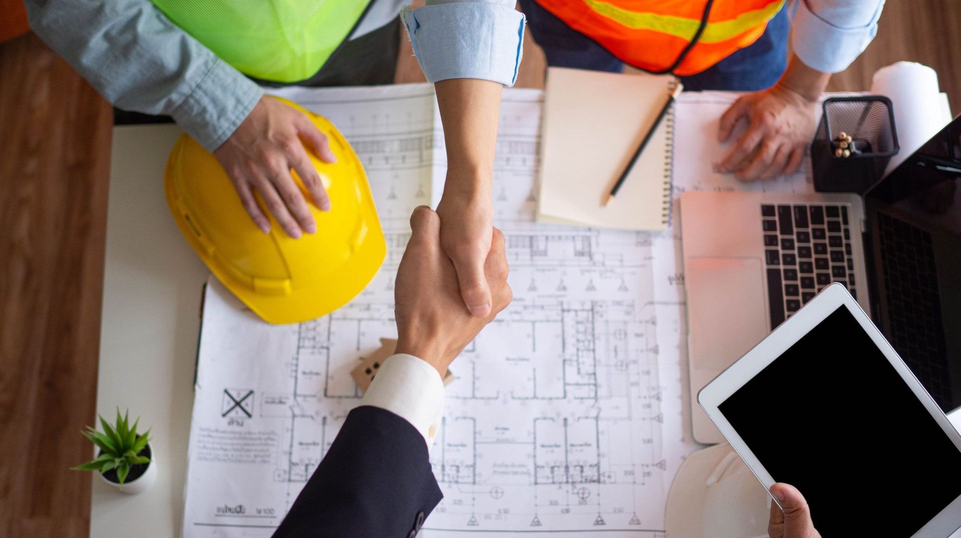 Two people shaking hands over architectural plans on a desk, one wearing a suit and the other in construction attire. Two people shaking hands over architectural plans on a desk, one wearing a suit and the other in construction attire.