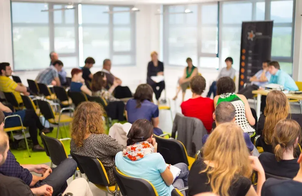 Audience at a panel discussion in a light-filled room; speakers in front; attendees in chairs, listening. Audience at a panel discussion in a light-filled room; speakers in front; attendees in chairs, listening.
