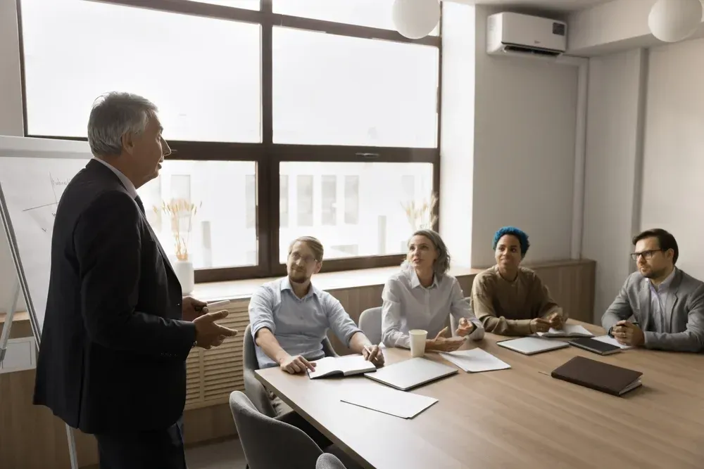 A businessman in suit giving a presentation to a diverse group seated at a conference table in a well-lit office. A businessman in suit giving a presentation to a diverse group seated at a conference table in a well-lit office.