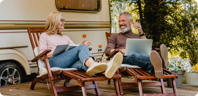 Couple relaxing in chairs, working on laptops next to an RV. Outdoors with trees. Smiling. Couple relaxing in chairs, working on laptops next to an RV. Outdoors with trees. Smiling.