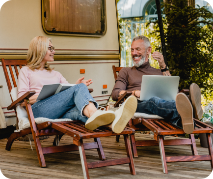 Couple relaxing on lounge chairs by a camper, woman holding a book, man with laptop, smiling.