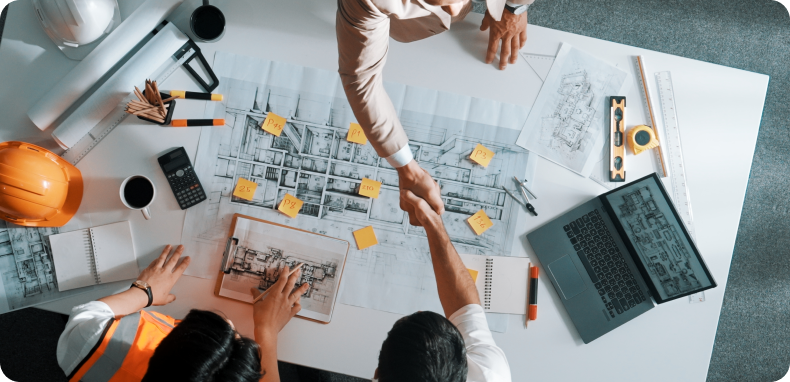 People shaking hands over blueprints and a laptop on a table. People shaking hands over blueprints and a laptop on a table.