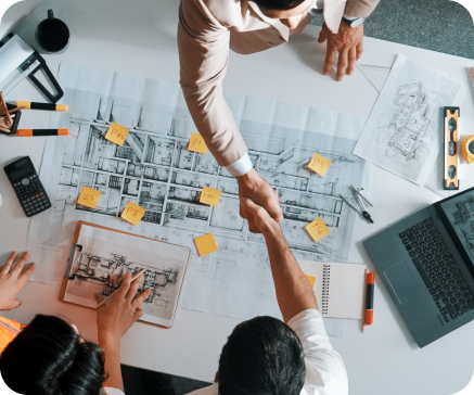 Architects shaking hands over blueprints, surrounded by tools and laptops.