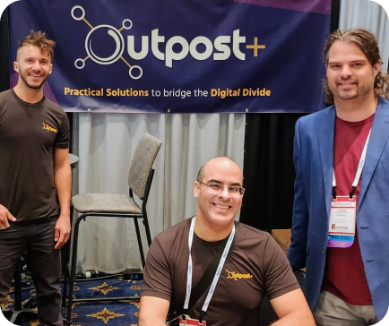 Three men at the Outpost+ booth. Banner says Three men at the Outpost+ booth. Banner says