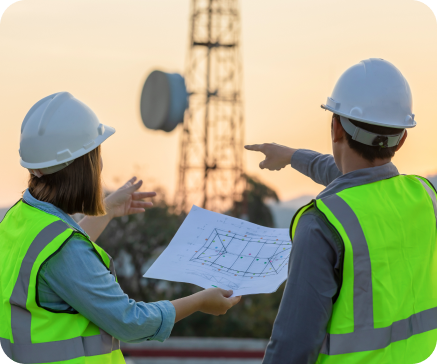 Two engineers in hard hats and vests review blueprints near a communication tower, pointing and discussing plans. Two engineers in hard hats and vests review blueprints near a communication tower, pointing and discussing plans.