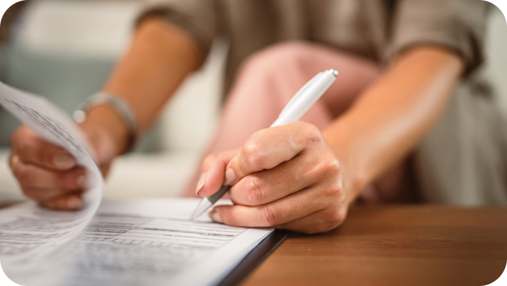 Woman writing on documents with a pen, seated at a table, close-up view.
