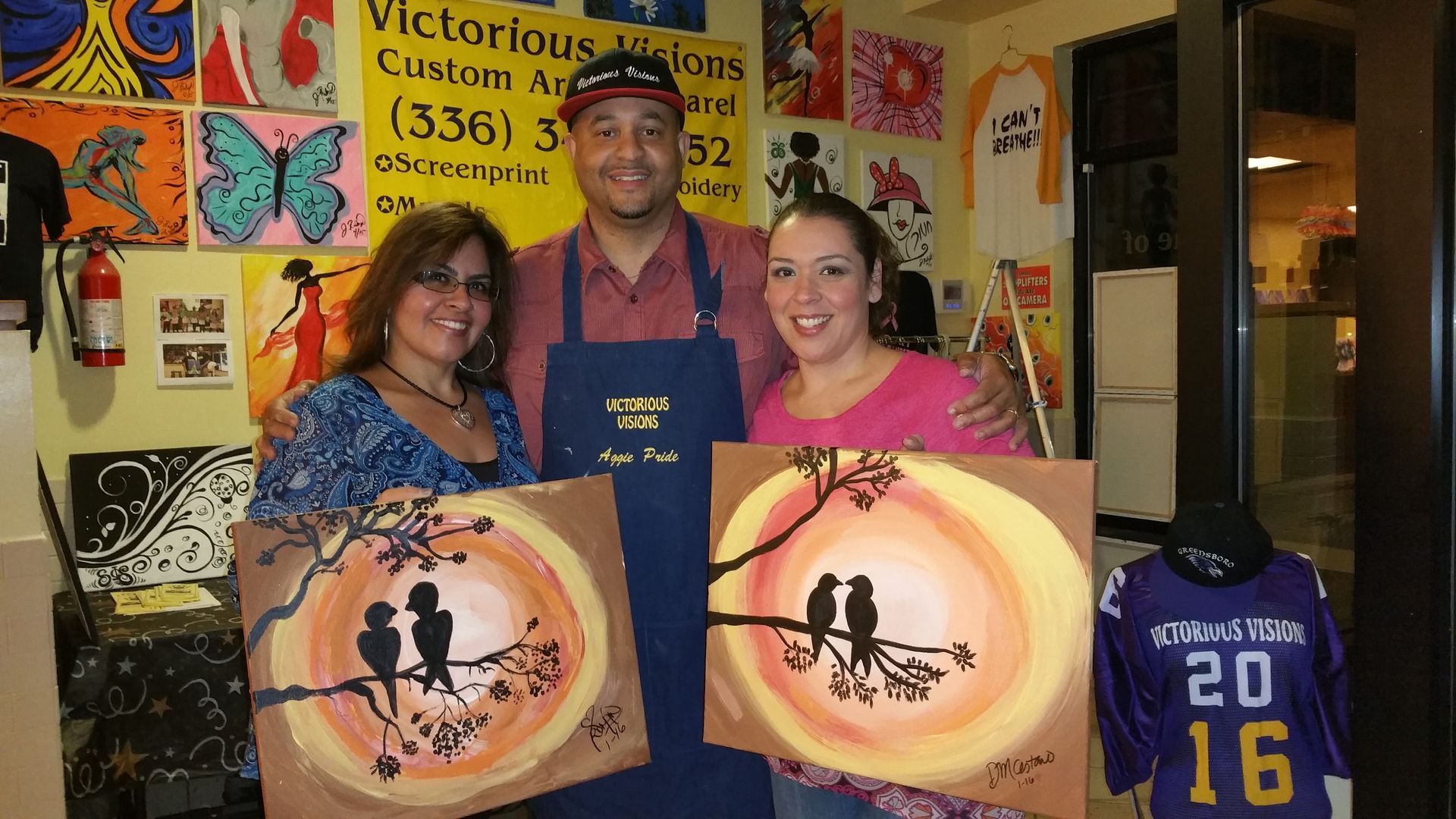 A man and two women holding paintings in front of a sign that says victorious creations