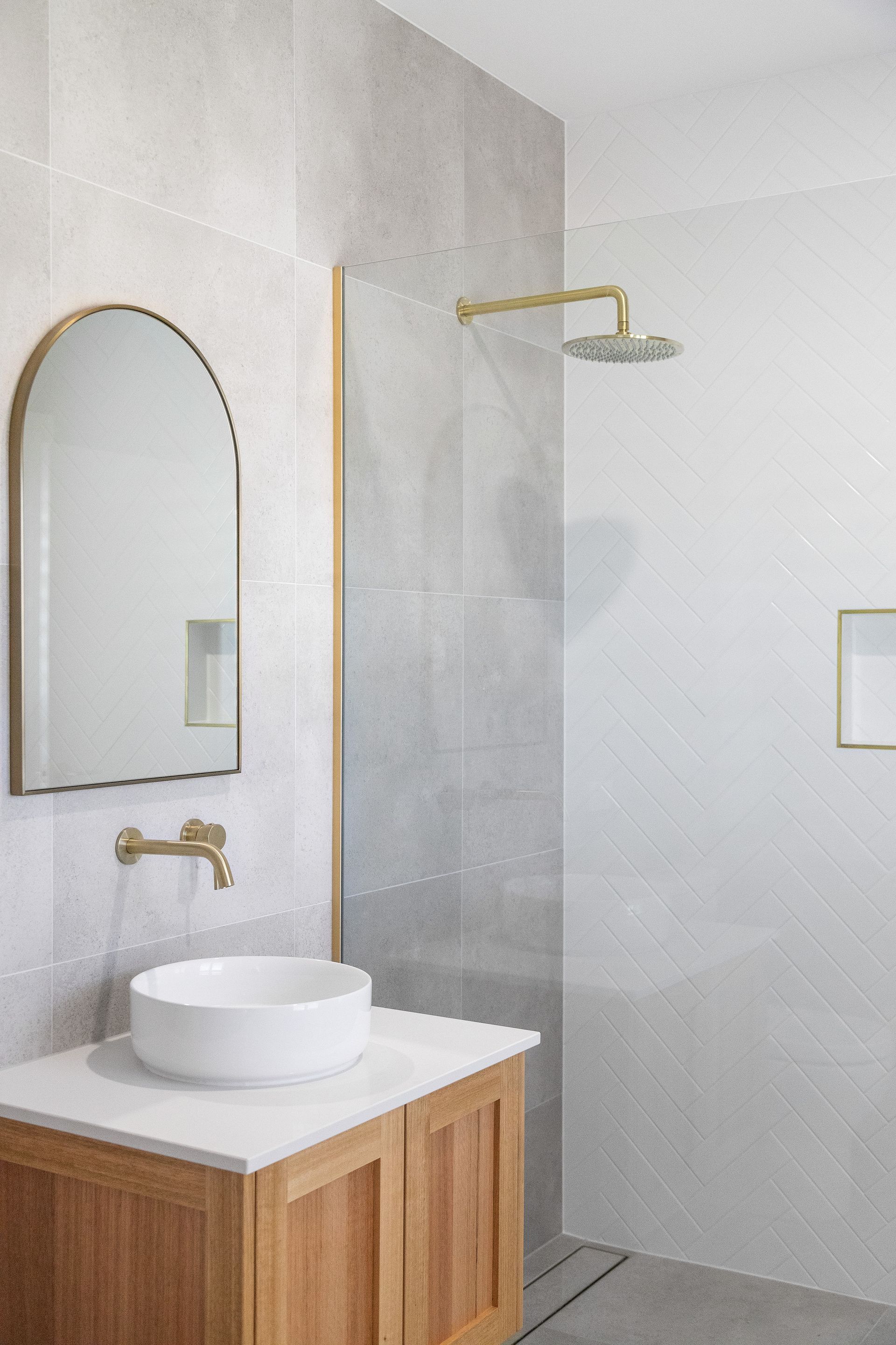 Modern bathroom with wooden vanity, round sink, gold faucet, arched mirror, and glass shower enclosure