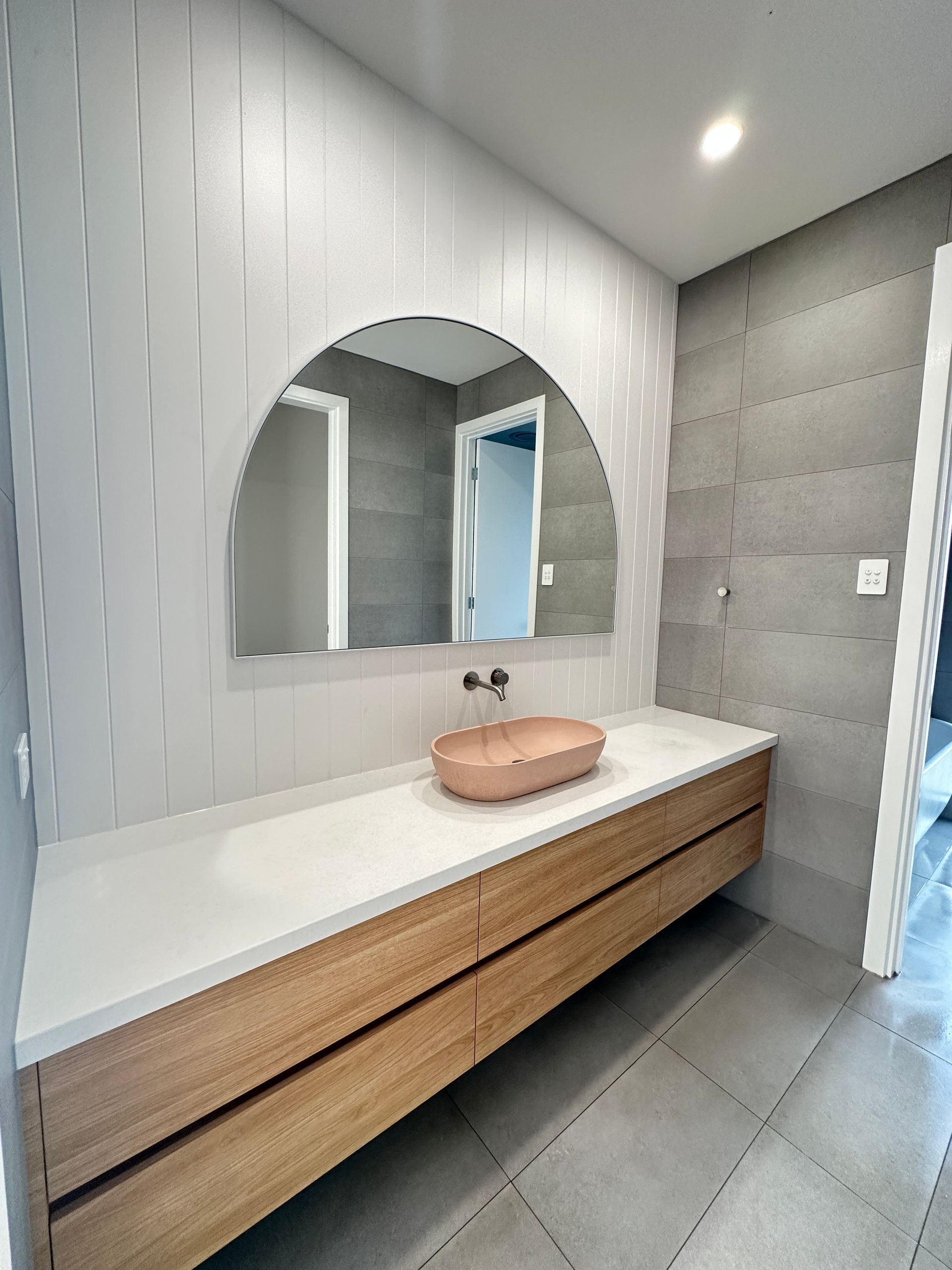 Modern bathroom vanity with round mirror, wood drawers, white countertop, and pink sink on gray tiled walls.