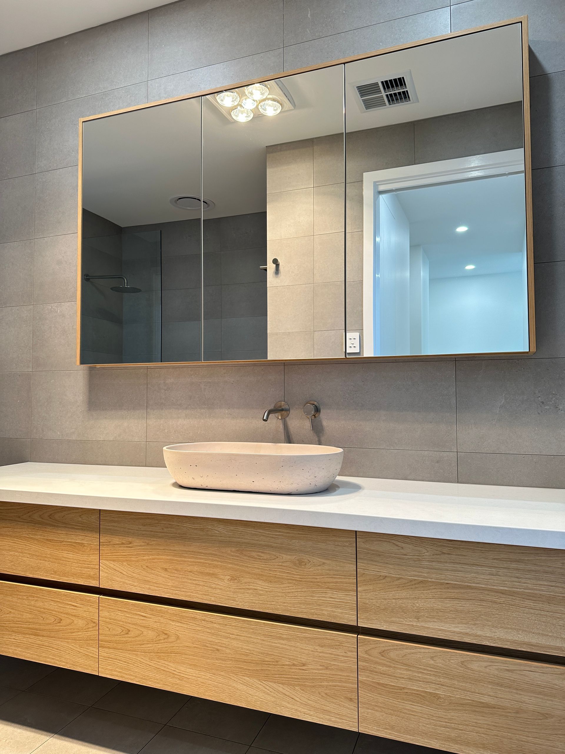Modern bathroom vanity with round sink, wall mirror, and light wood cabinet against a gray stone wall