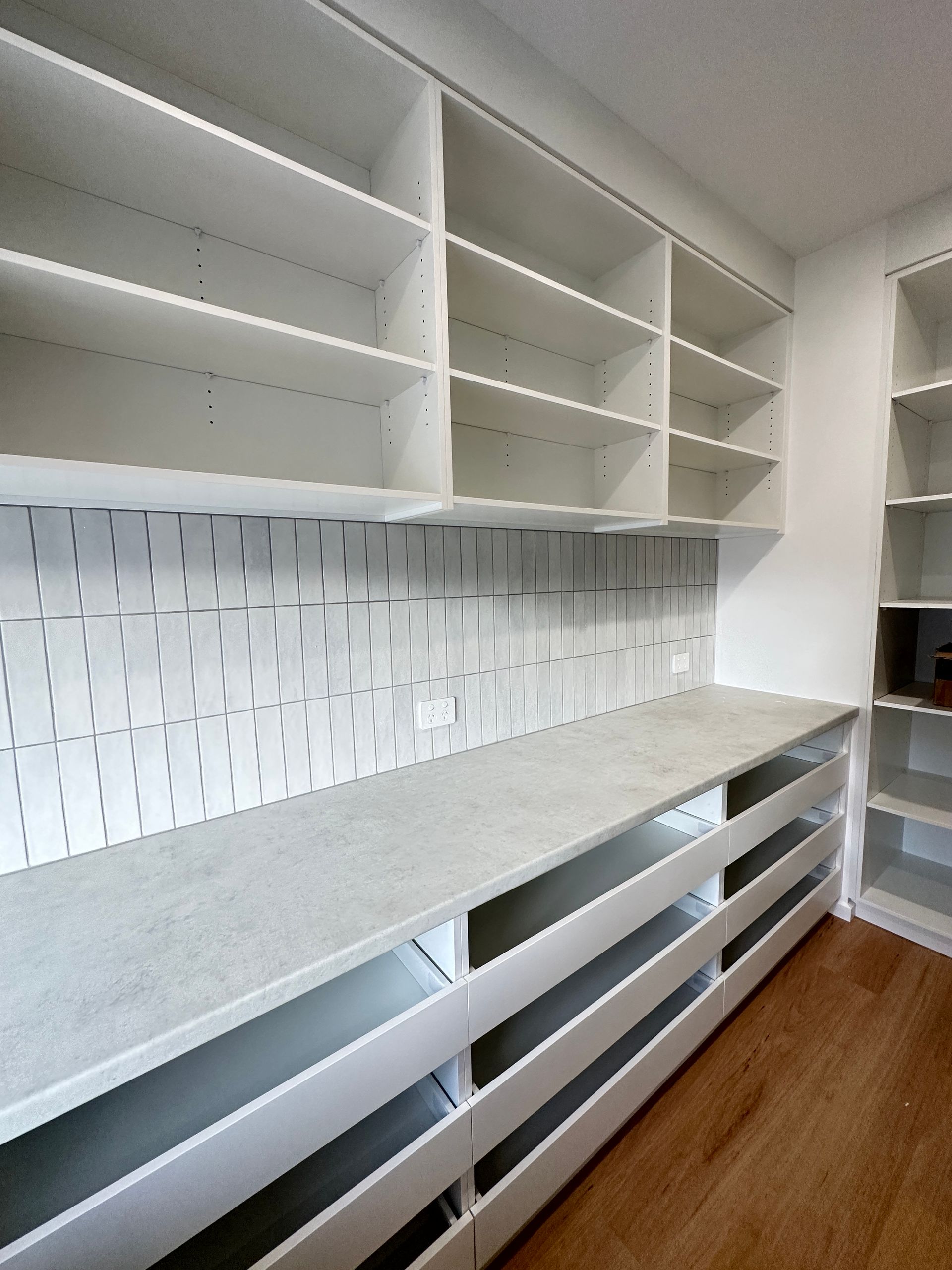 White kitchen with open shelves, gray countertops, and drawers along a tiled backsplash and wood floor.