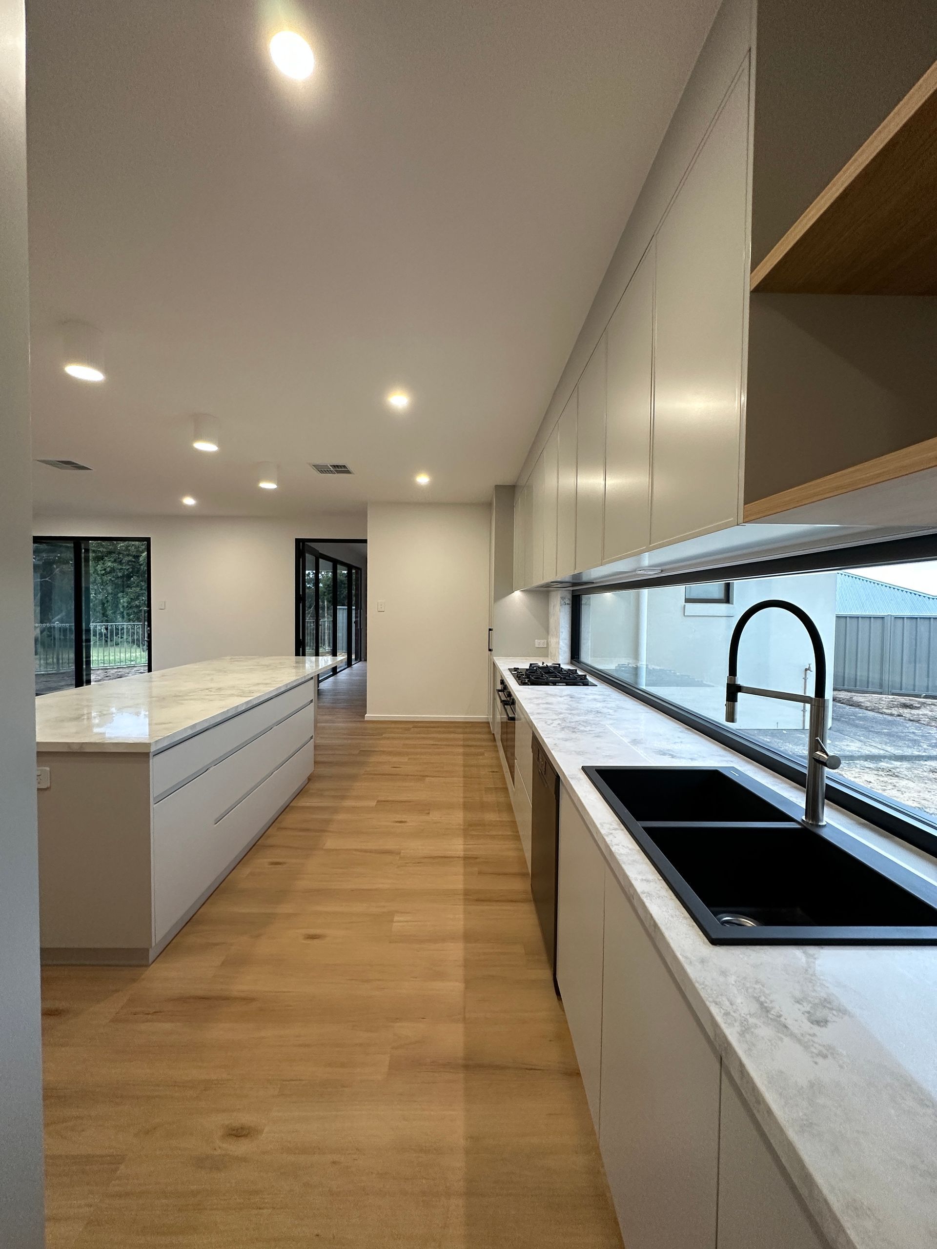 Modern white kitchen with wood floors, long island, and large window over a black sink and marble countertop