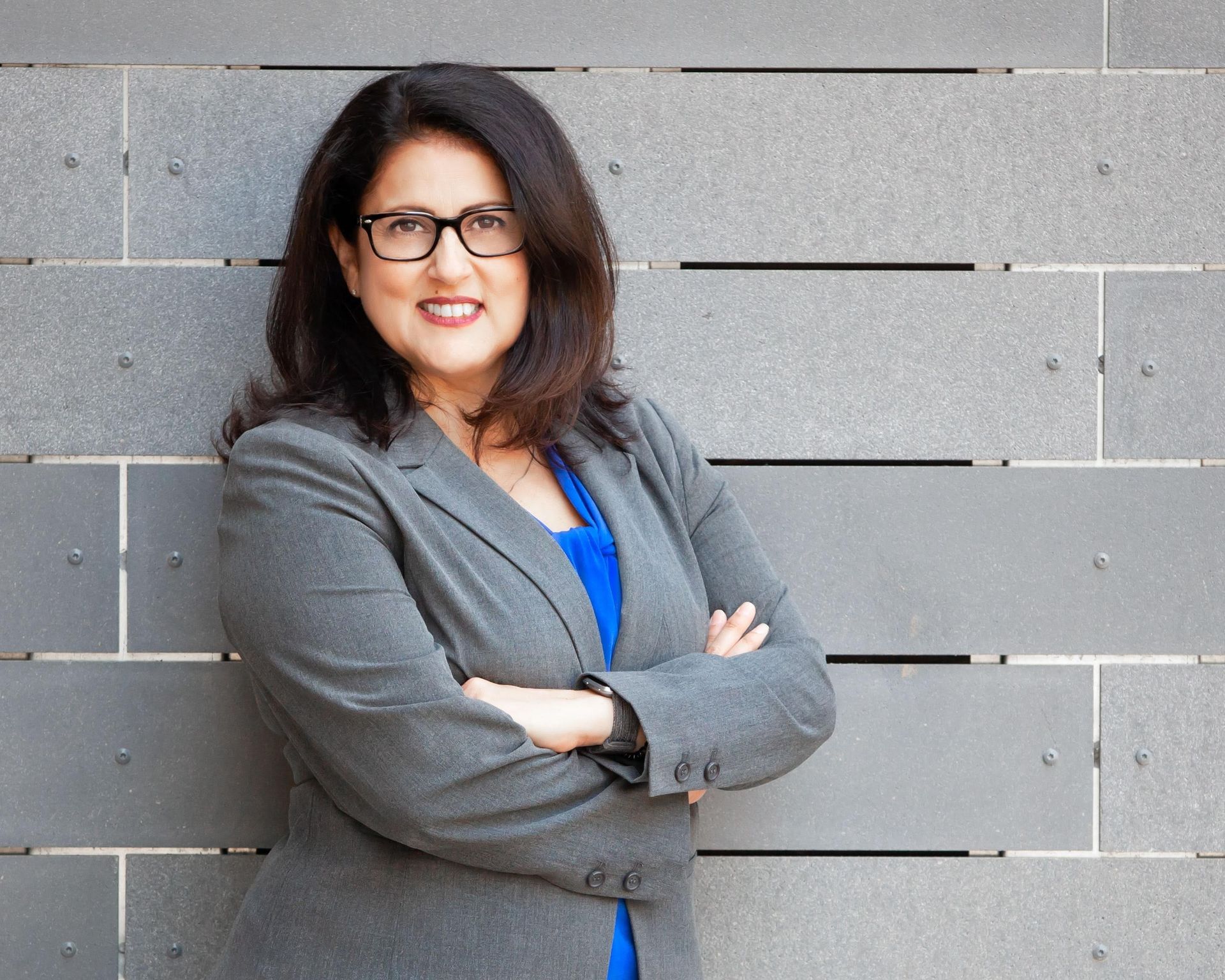 A woman in a suit and glasses is standing in front of a brick wall with her arms crossed.