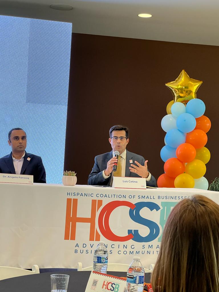 A man speaking into a microphone in front of a sign that says hcs