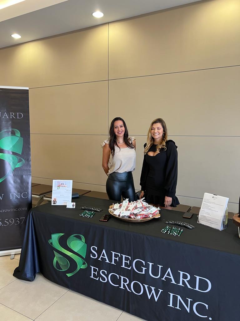 Two women are standing at a table with a safeguard escrow inc. table cloth.