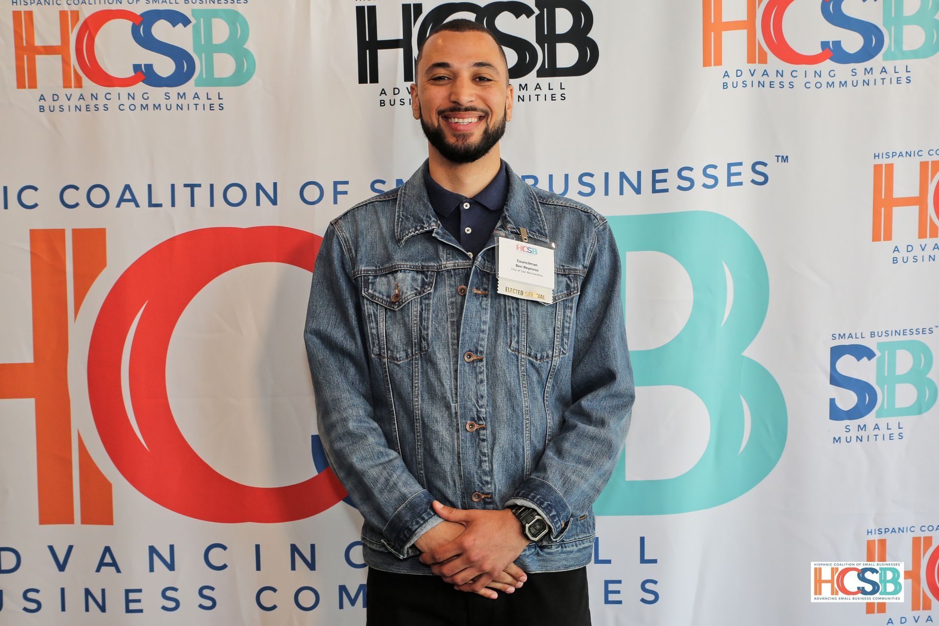 A man in a denim jacket is standing in front of a hcsb banner