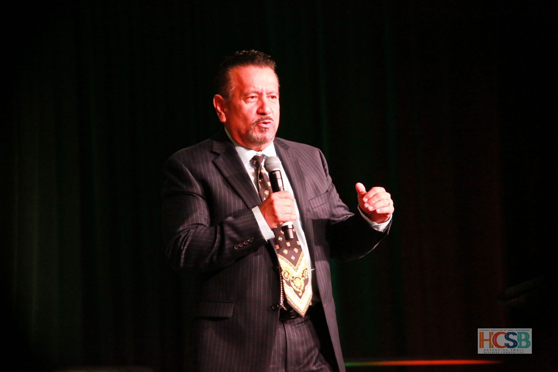 A man in a suit and tie is speaking into a microphone on a stage.