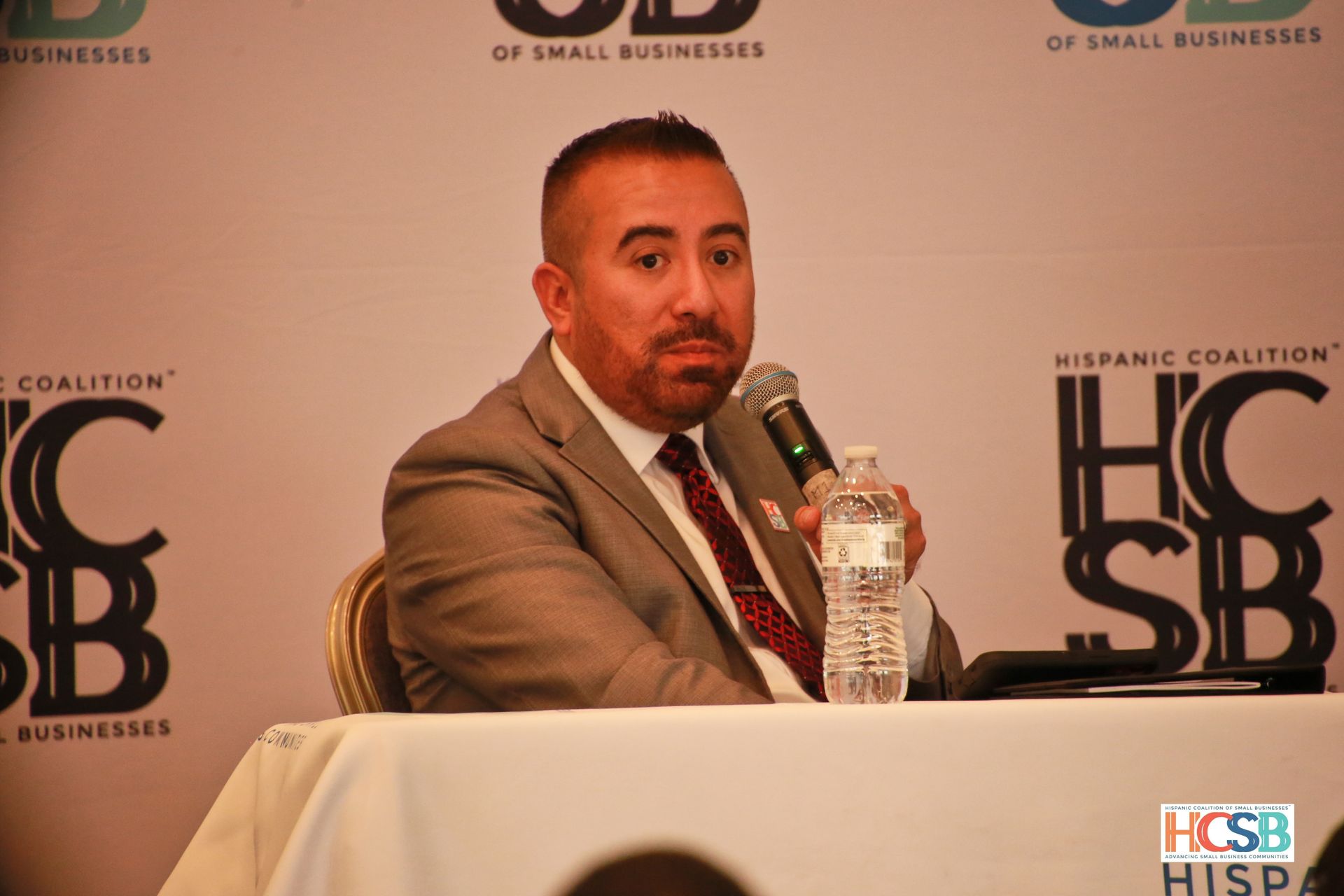 A man in a suit and tie is sitting at a table holding a microphone and a bottle of water.
