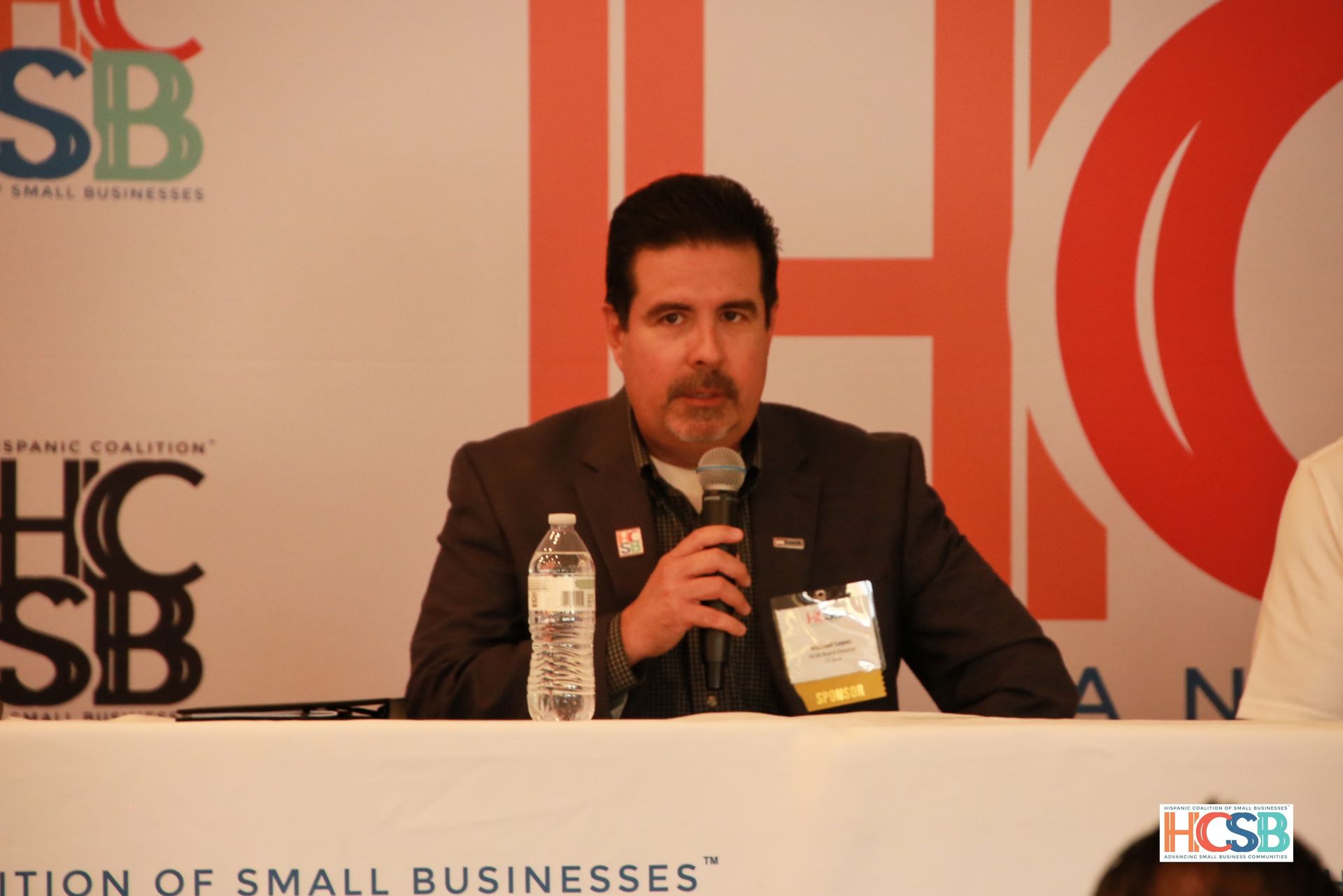 A man sitting at a table with a microphone in front of a sign that says sb